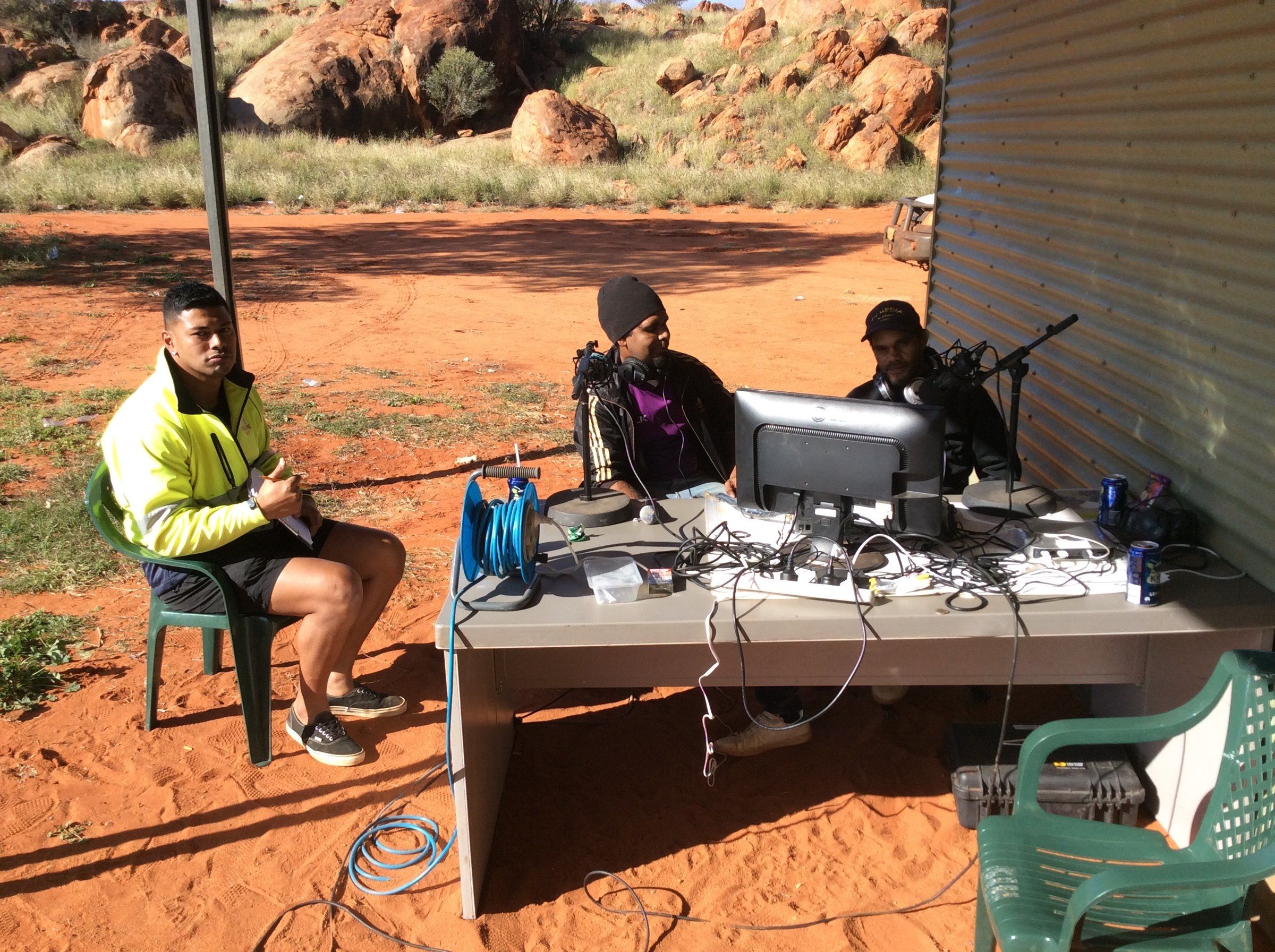 An outside broadcast set up on red earth beside a community building, with the radio desk in the open air